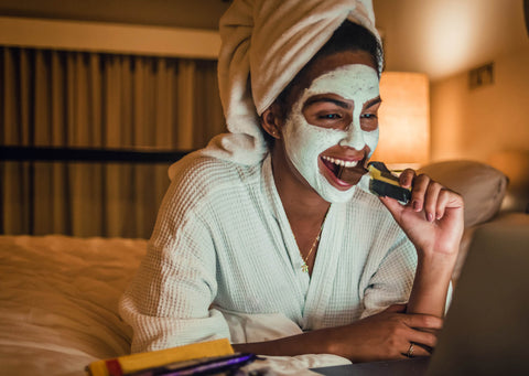 Frau im Bademantel mit Handtuchturban und Gesichtsmaske sitzt entspannt auf dem Bett, beißt in eine Schokoladentafel und schaut lächelnd auf ihren Laptop.
