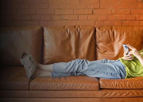 Eine Person liegt entspannt mit überschlagenen Beinen auf der Couch und schaut auf ihr Smartphone.